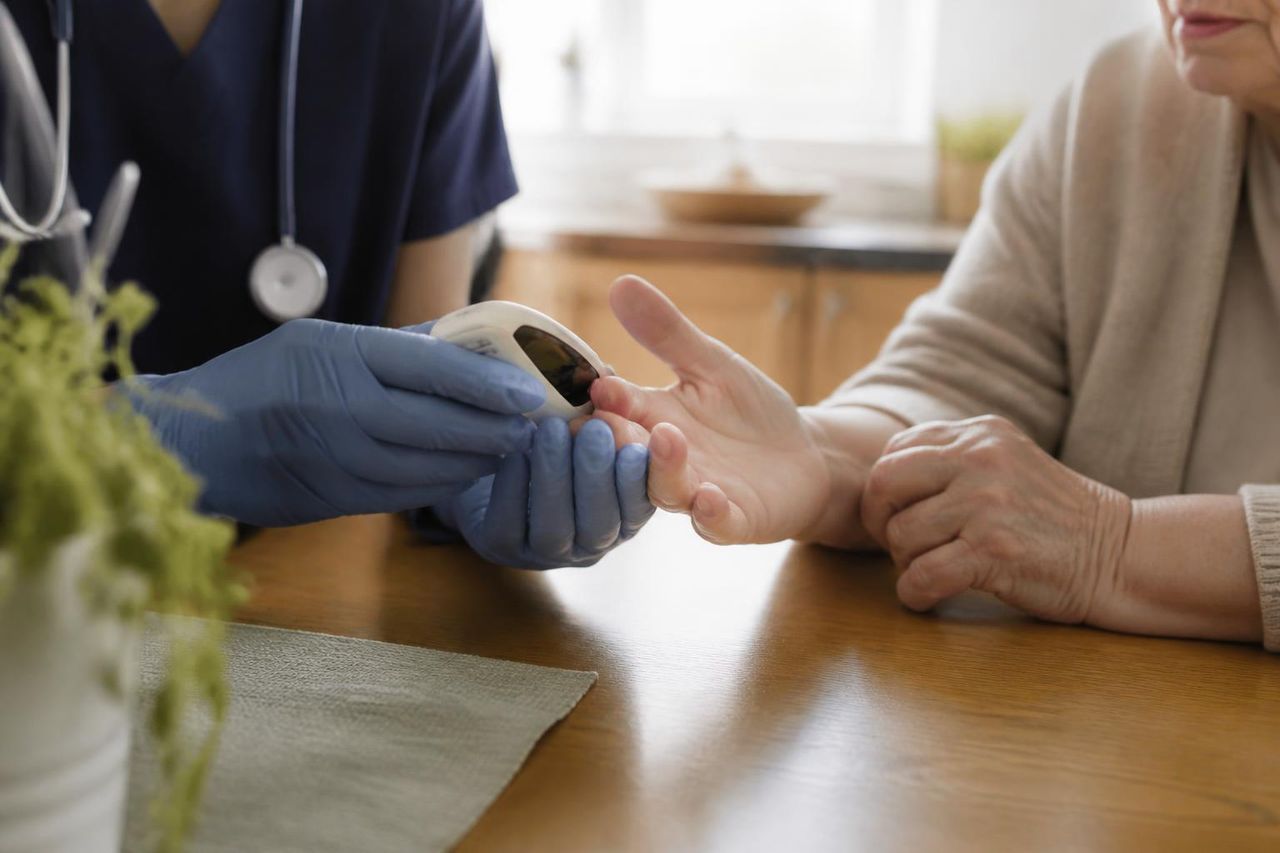 Gloved nurse hands using a small handheld glucose monitor beside an older person's hand on a kitchen table