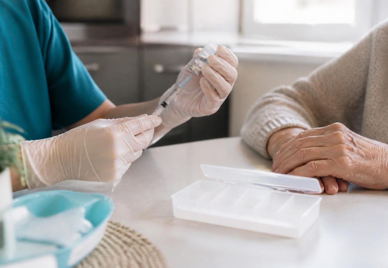 Gloved nurse hands drawing up a prescribed injection beside a weekly pill organiser on a home bench